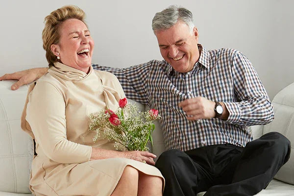 Happy senior couple sitting on sofa, woman holding flowers, enjoying comfortable adaptive clothing
