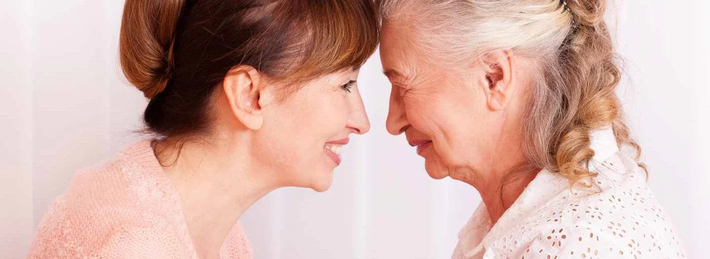 Smiling senior woman and caregiver share a supportive moment indoors, representing adaptive clothing and dementia care.