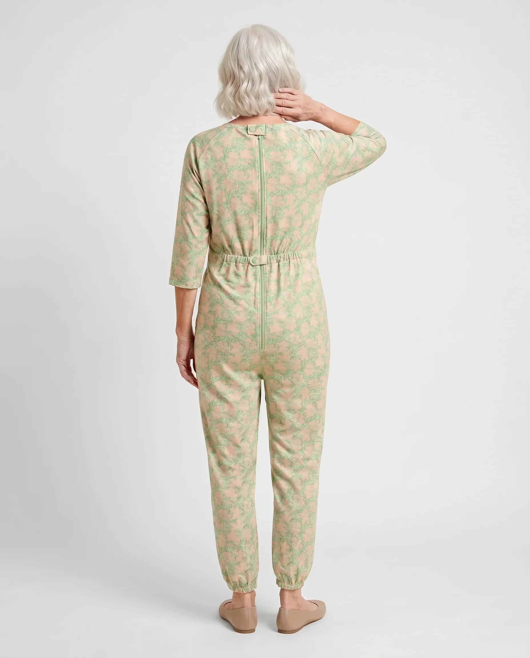 Senior woman wearing Ovidis adaptive Carrie Apple Green floral jumpsuit, full body back view showing center-back zipper and snap closures