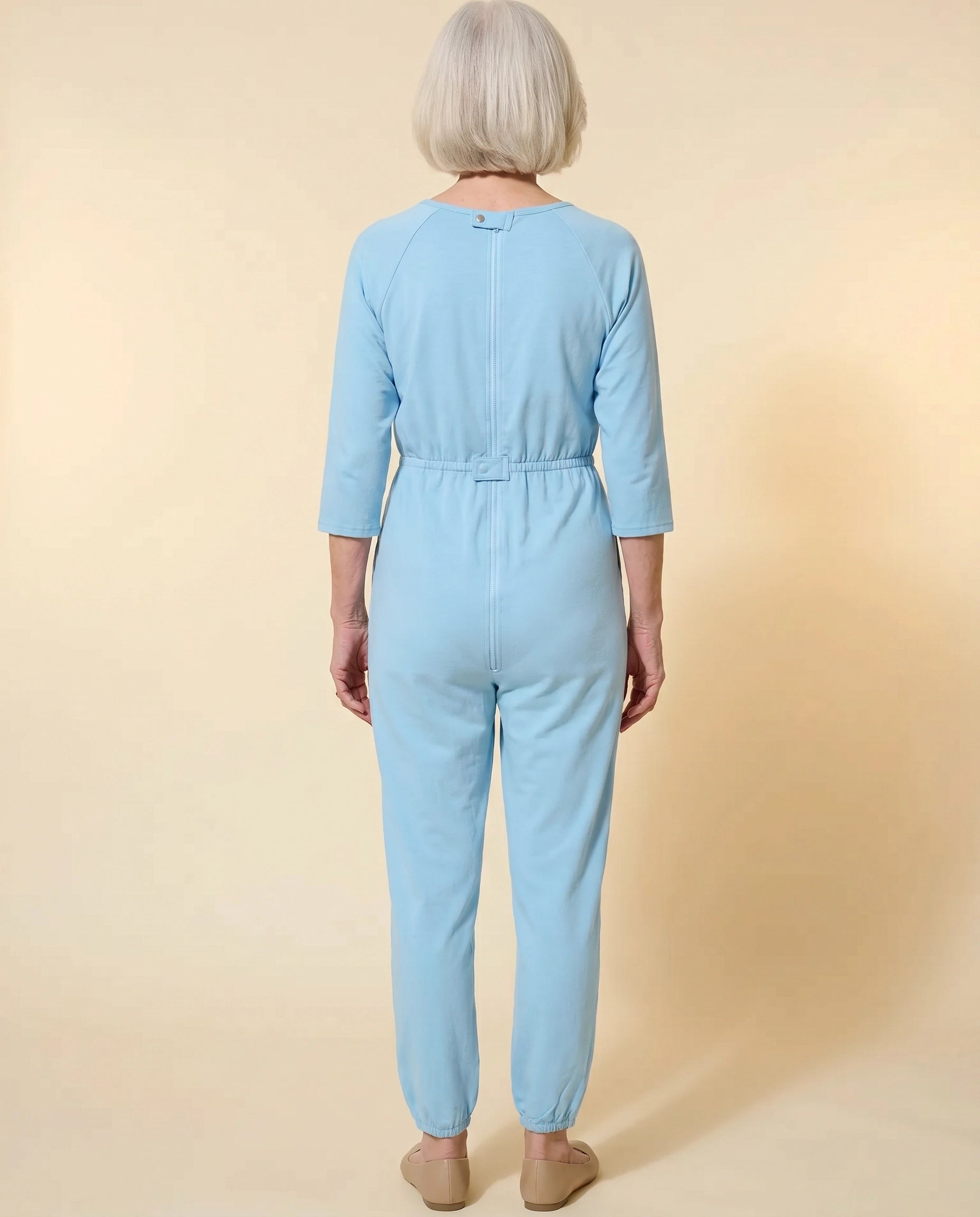 Senior woman wearing Ovidis adaptive Carrie Blue jumpsuit, full body back view showing center-back zipper and snap closures