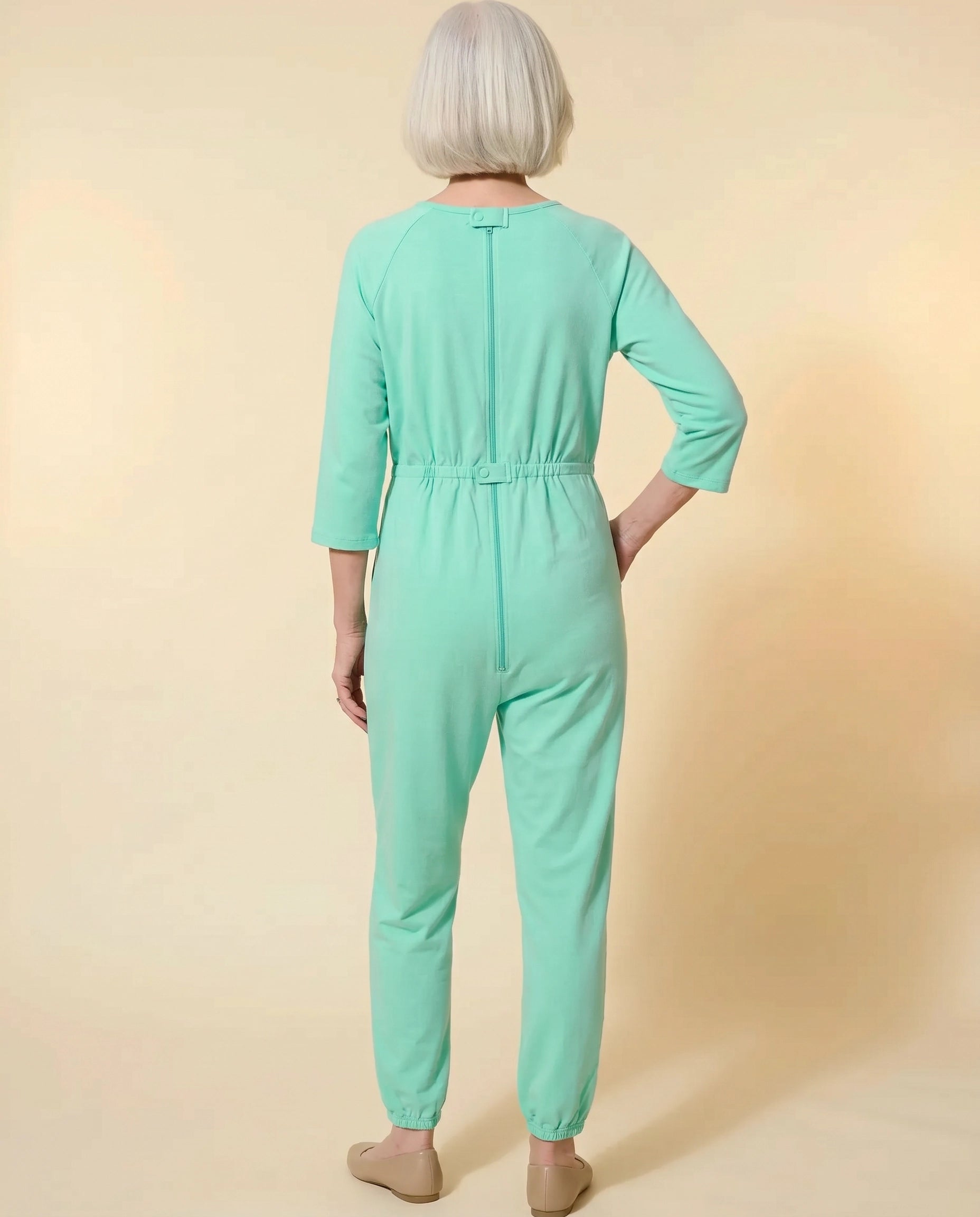 Senior woman wearing Ovidis adaptive Carrie Green jumpsuit, full body back view showing center-back zipper and snap closures