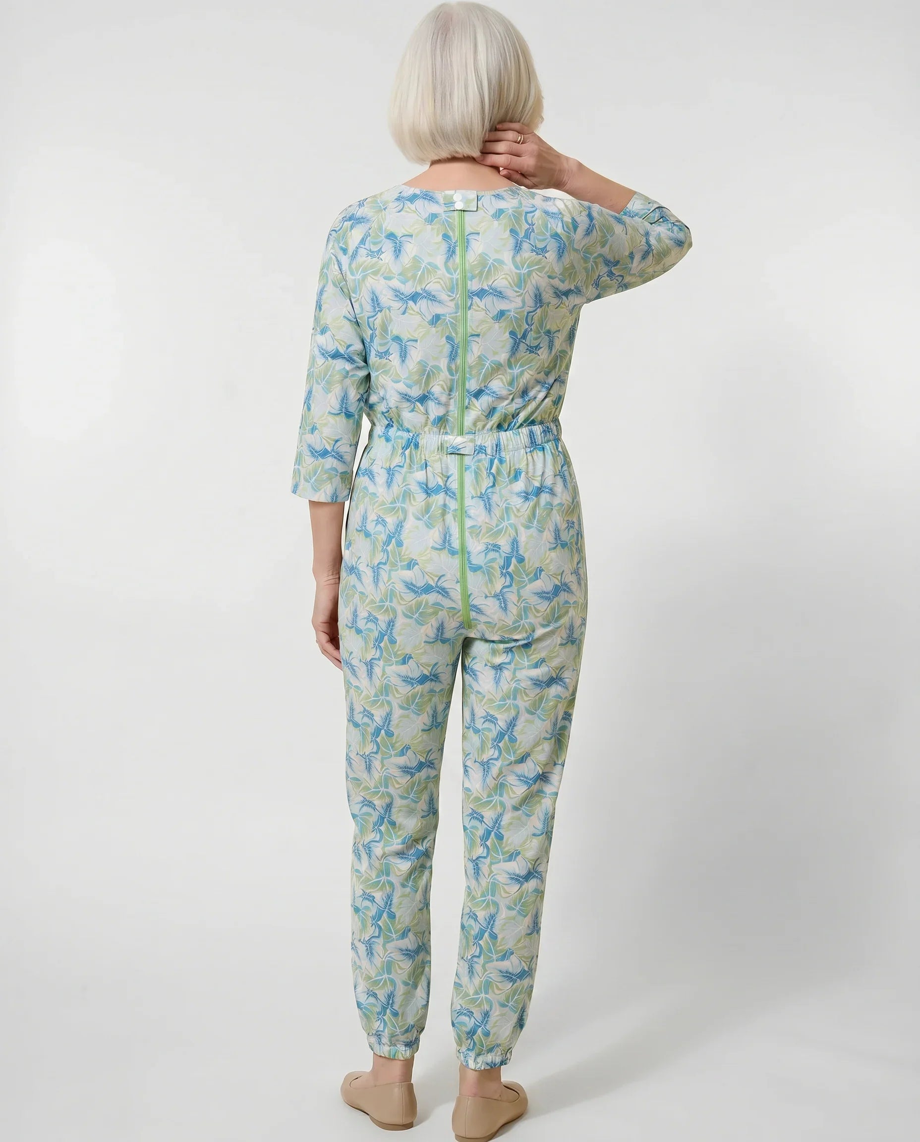 Senior woman wearing Ovidis adaptive Carrie Mint tropical leaf jumpsuit, full body back view showing center-back zipper and snap closures