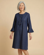 Senior Asian woman wearing Loulou adaptive denim dress with ruffle placket and bell sleeves, parchment backdrop