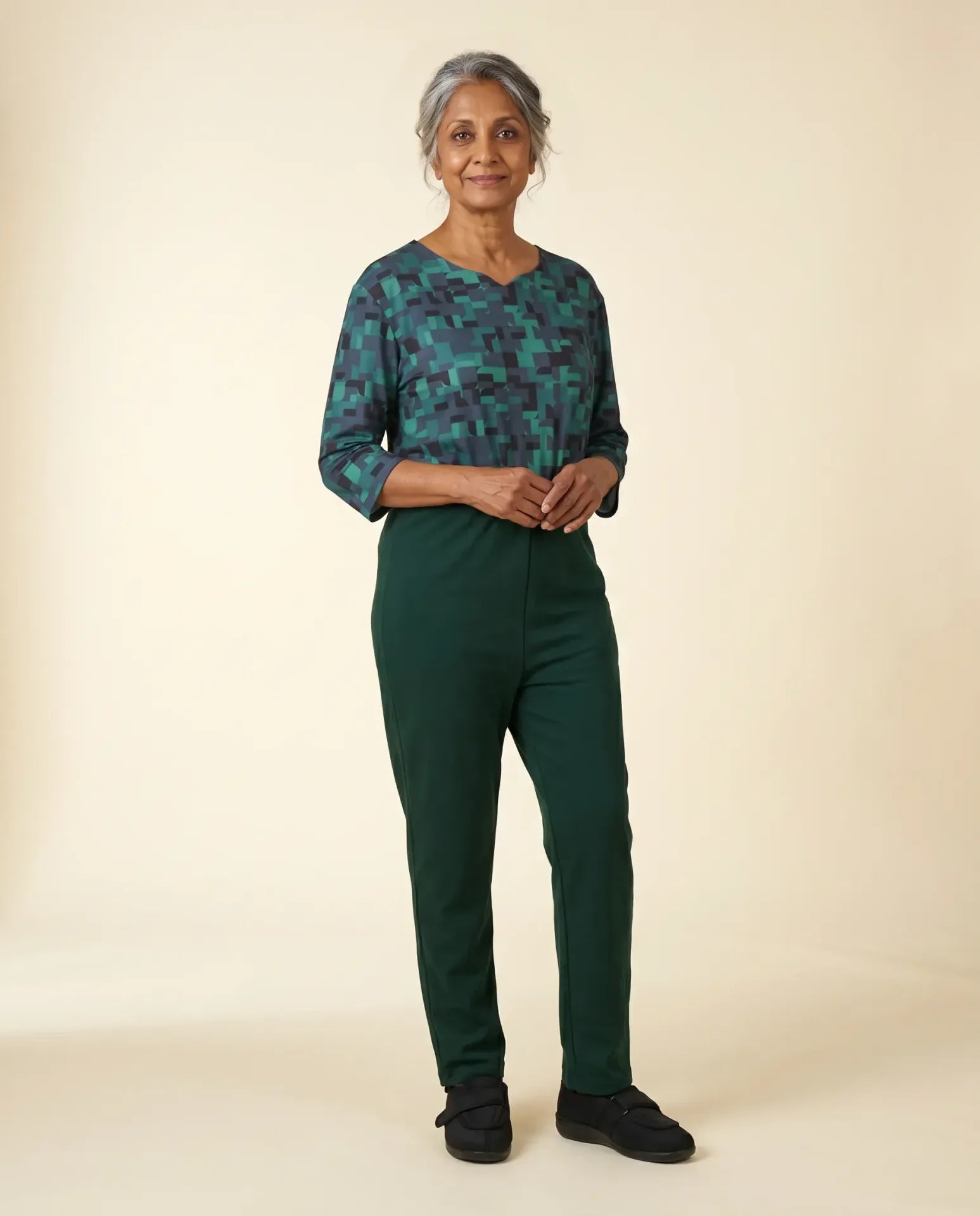 Senior woman wearing Ovidis adaptive Bella Hummingbird jumpsuit — teal pixel print with forest green pants